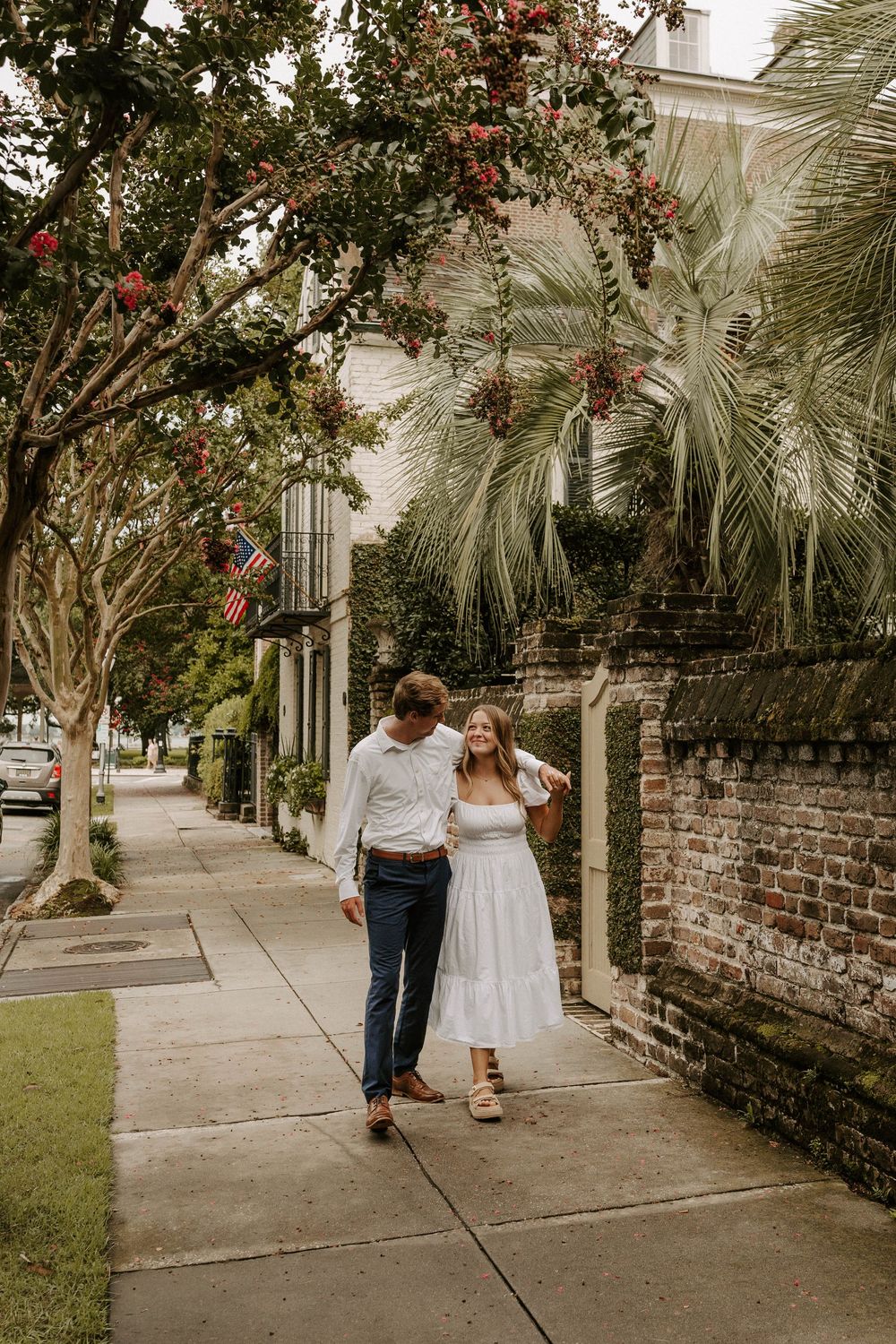 Couple walking hand in hand on a charming brick-lined sidewalk with palm trees and American flags in background.