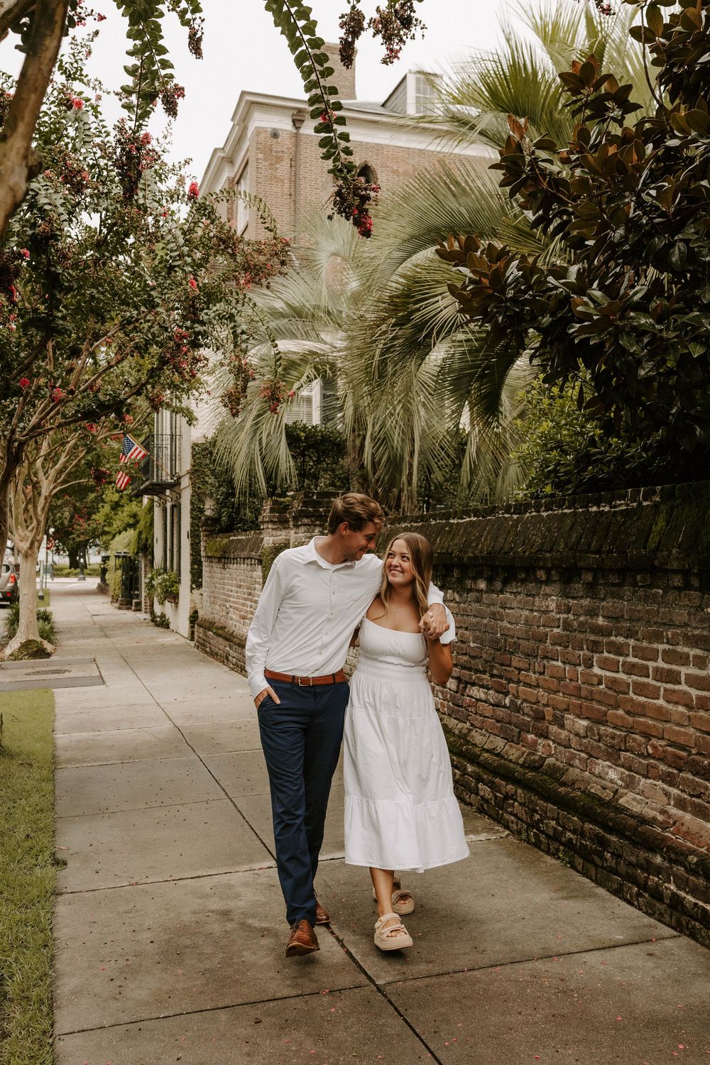 Couple walking hand in hand on a charming brick-walled sidewalk lined with lush greenery and palm trees.