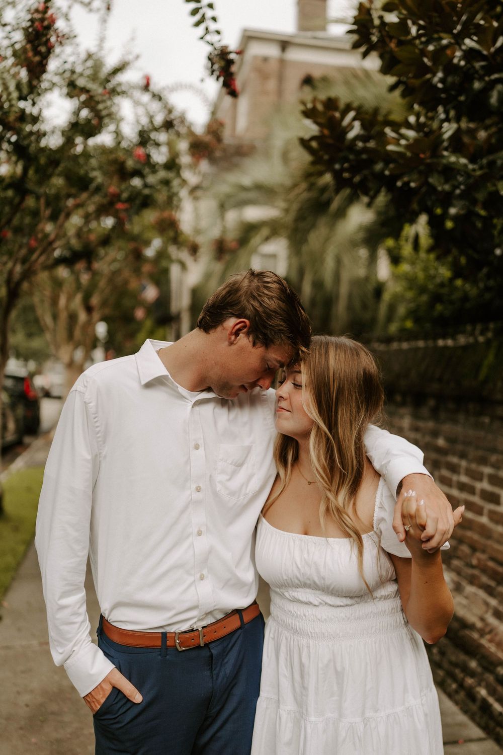 Couple in matching white outfits share a tender moment walking along a tree-lined street.