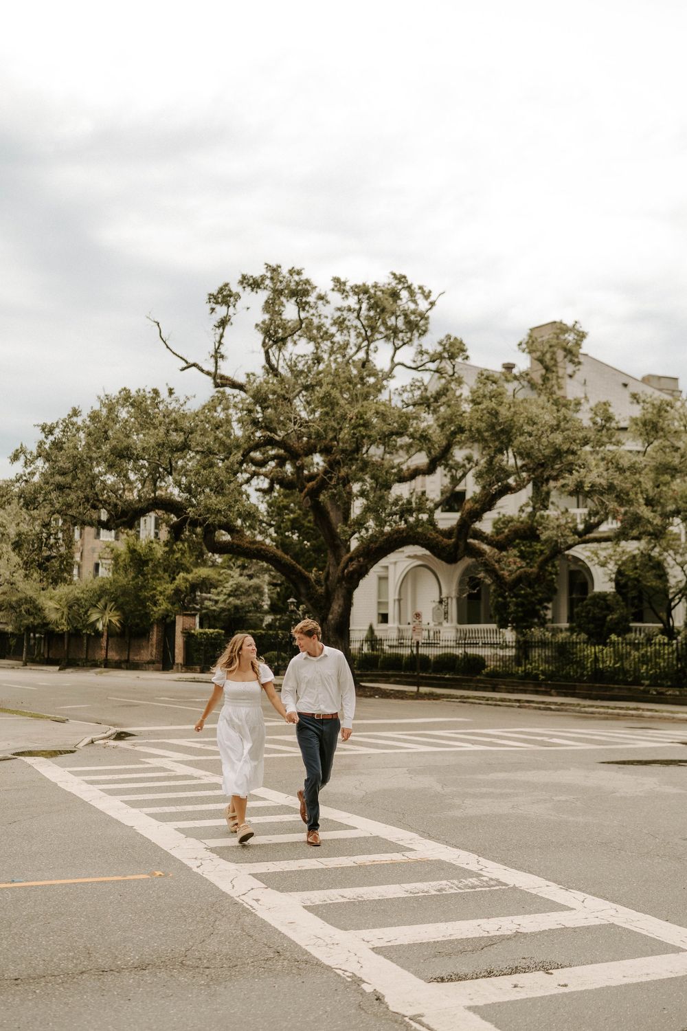 Couple holding hands crossing a street crosswalk with a large oak tree and white building behind them.