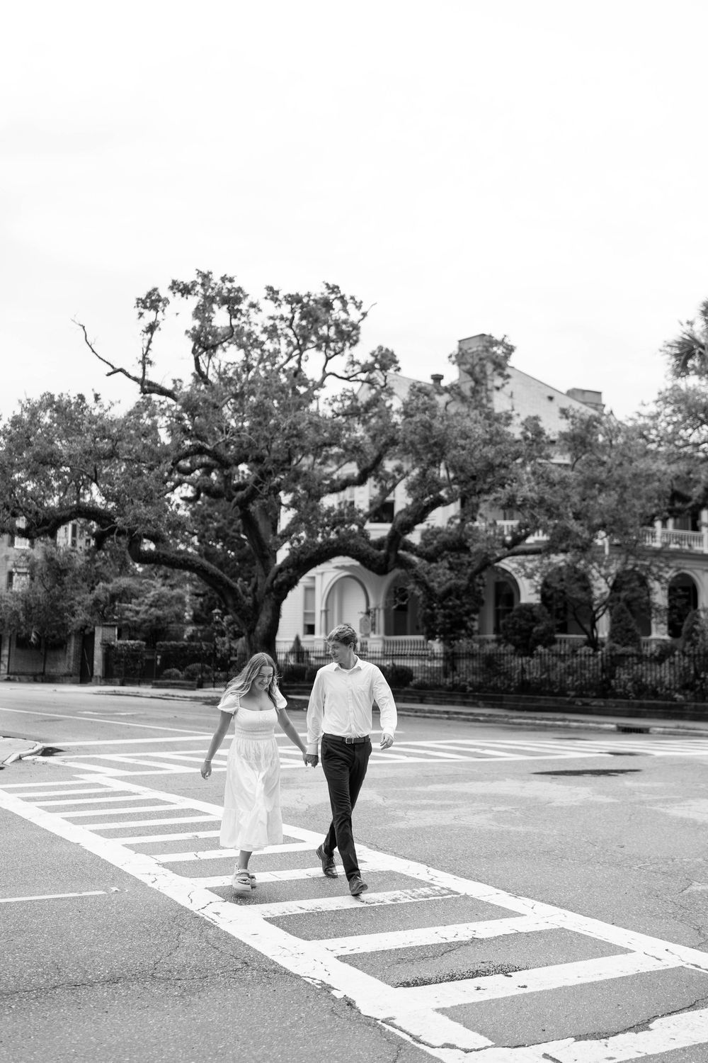 Black and white photo of a couple holding hands while crossing a street crosswalk with a large oak tree behind them.