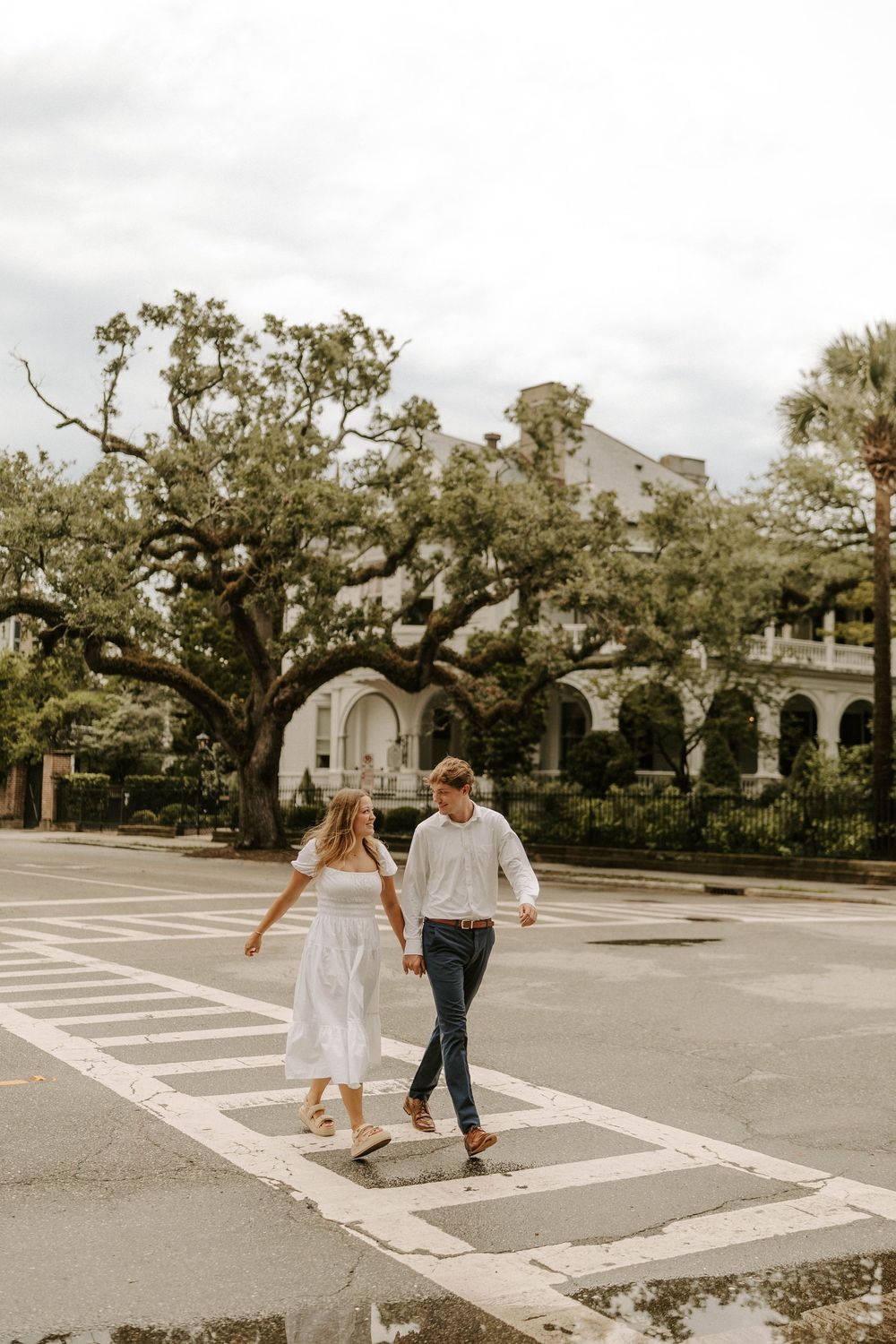 Happy couple holding hands crossing a street with a grand oak tree and historic building behind.