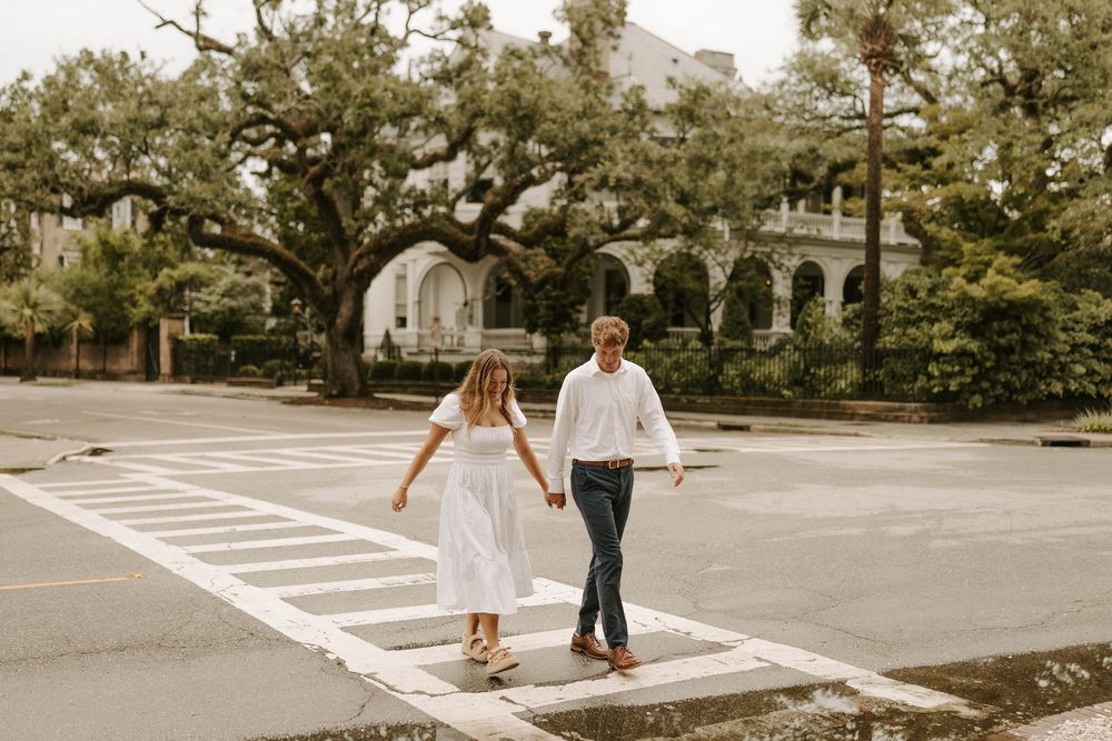 Couple holding hands crossing a zebra crossing on a tree-lined street, dressed in white outfits on a cloudy day.