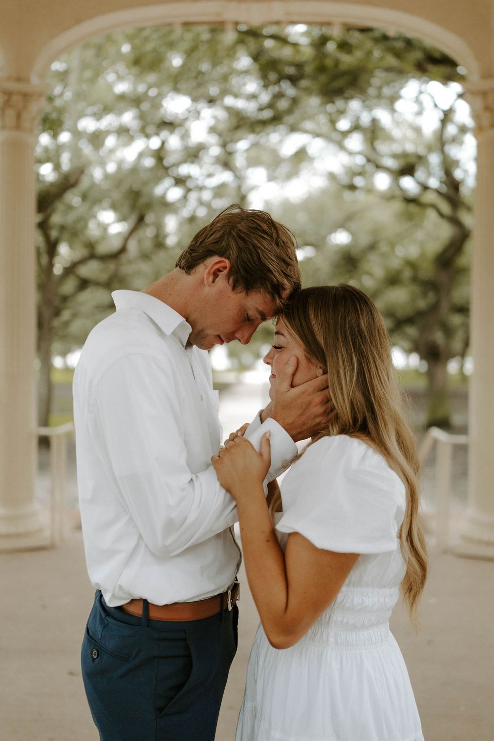 Romantic couple in white and navy touching foreheads under outdoor pavilion.