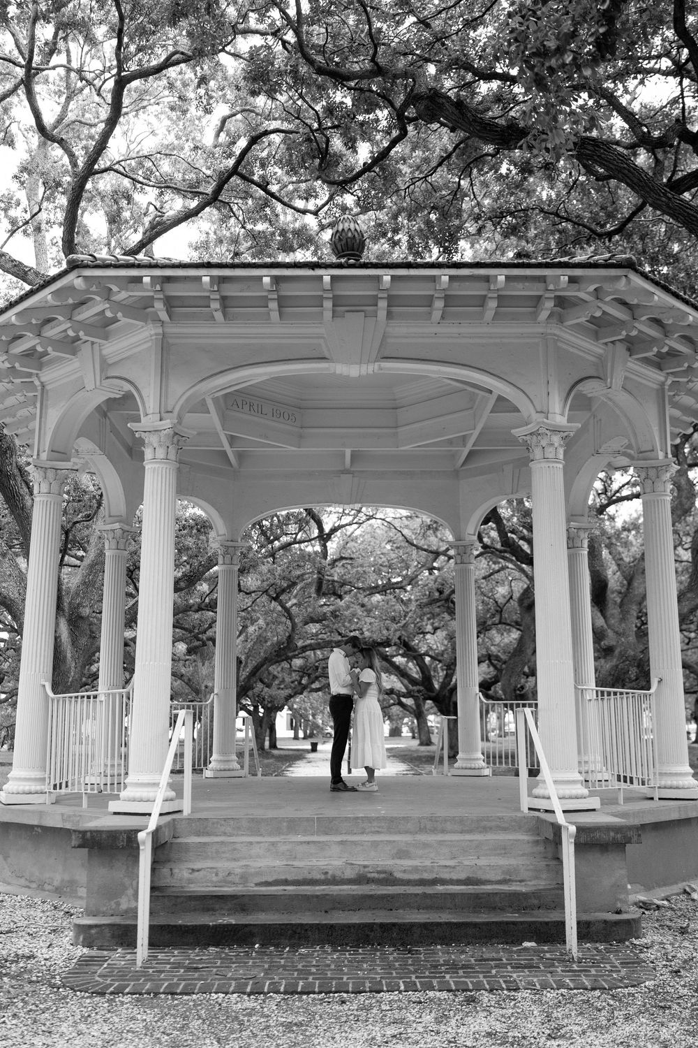 Couple shares a romantic moment in a white gazebo surrounded by oak trees in a park.