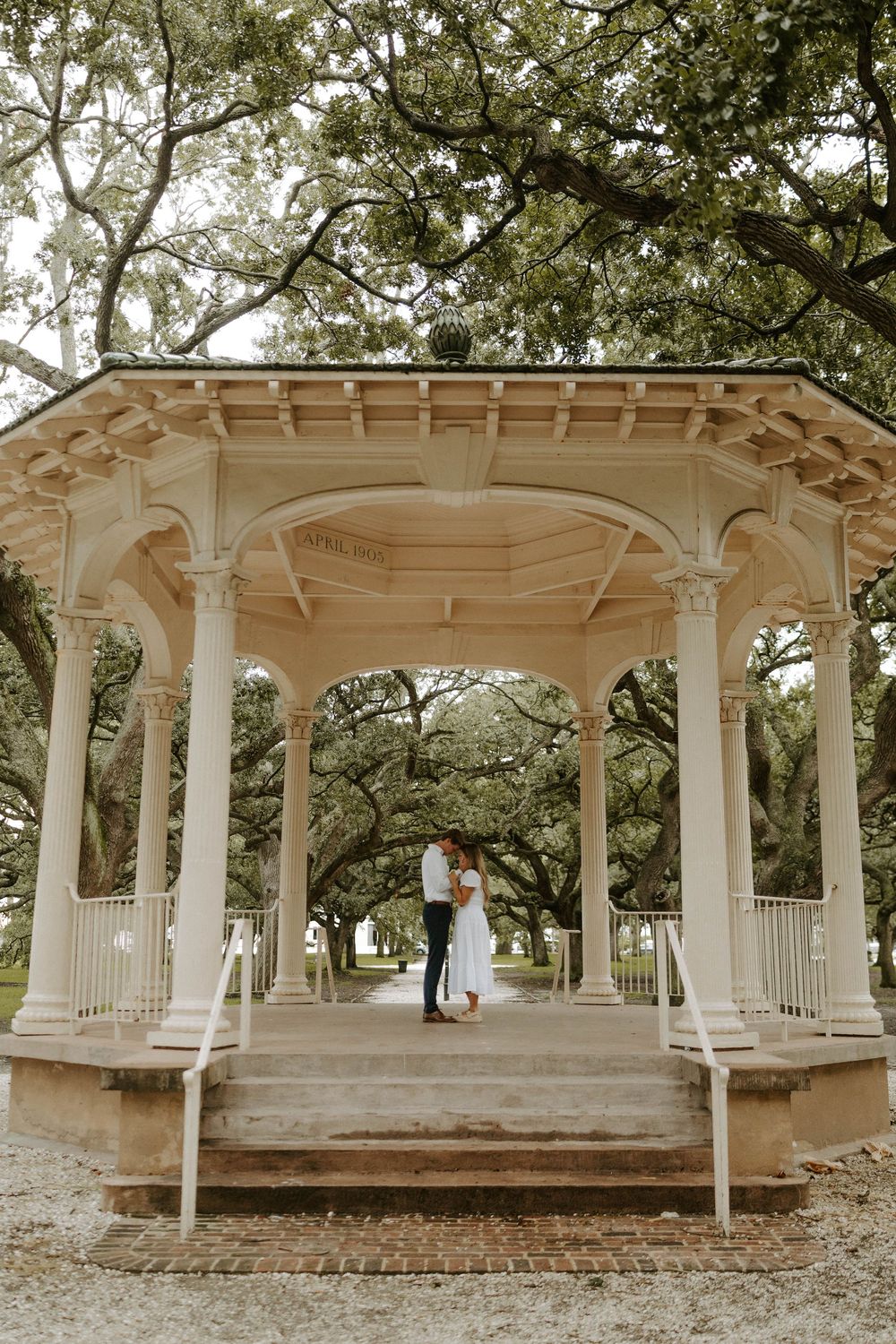 Couple embracing under elegant white gazebo surrounded by lush oak trees in a romantic outdoor setting.