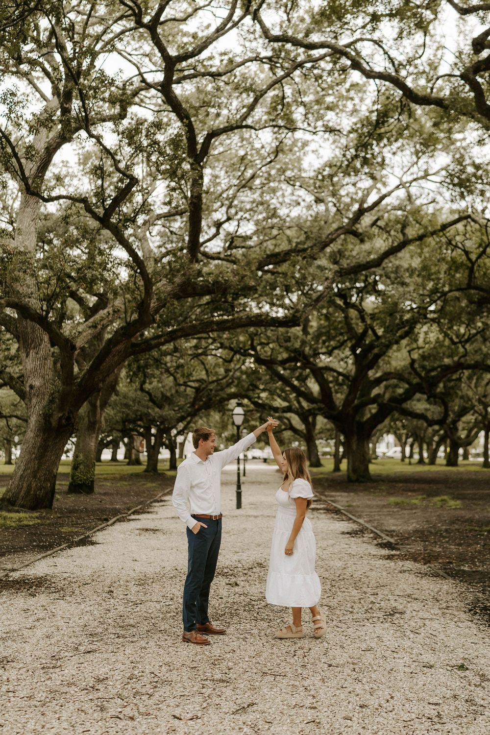A couple dances on a gravel path surrounded by sprawling oak trees, the woman wearing a white dress.