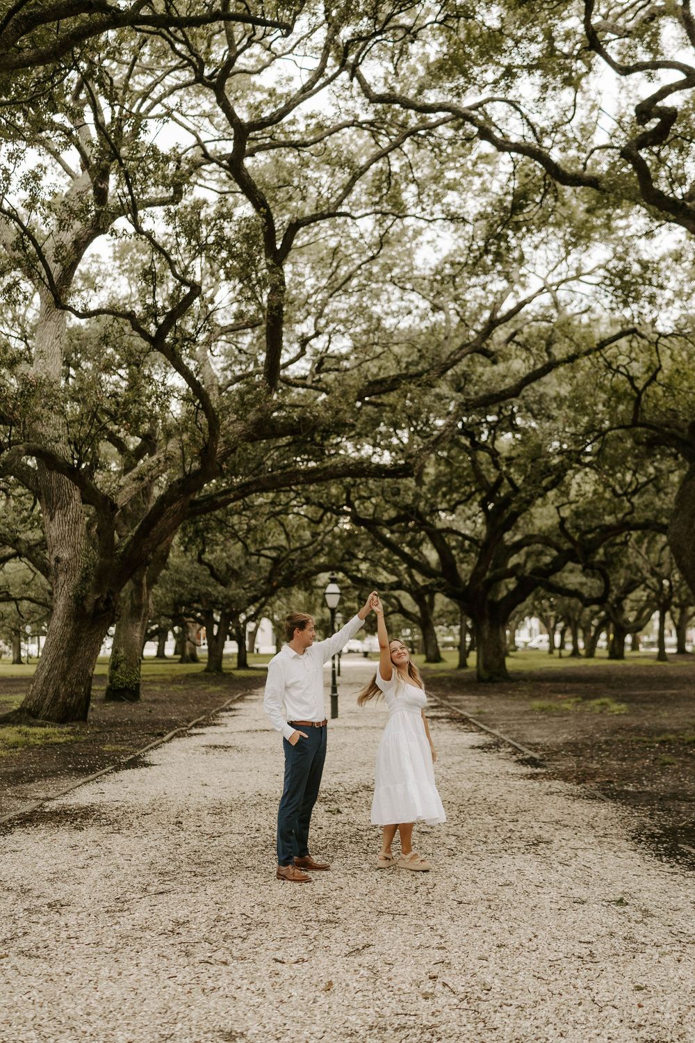 Couple dancing under sprawling oak trees on a gravel path, woman in white dress, man in white shirt and jeans.