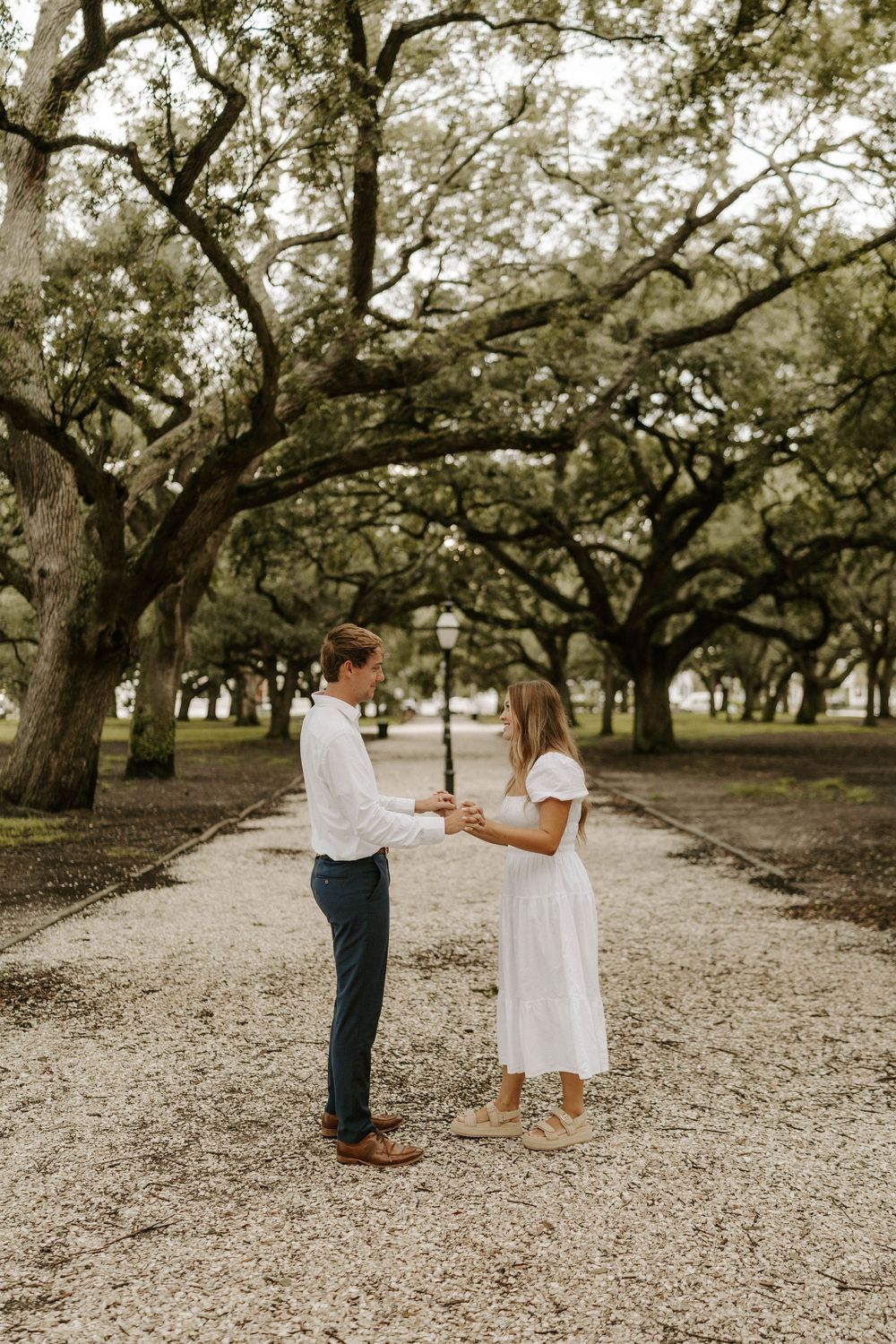 Couple in white outfits holding hands on a tree-lined gravel path during an engagement photo session.