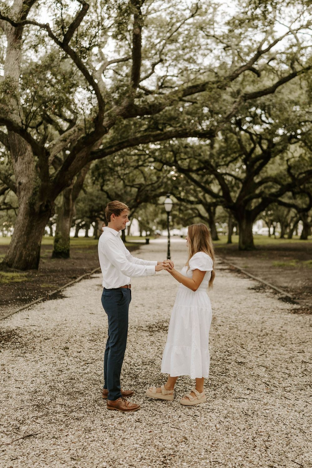 Couple holds hands on a tree-lined gravel path during an outdoor engagement session.