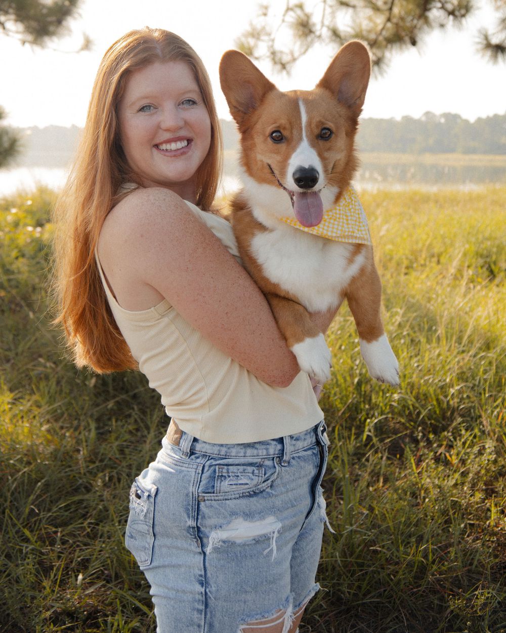 Smiling young woman with red hair holding an adorable Corgi puppy wearing a yellow bandana outdoors.