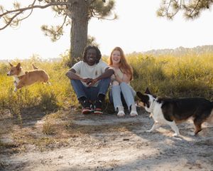 Happy couple sitting outdoors with two playful Corgis in a sunny natural setting.
