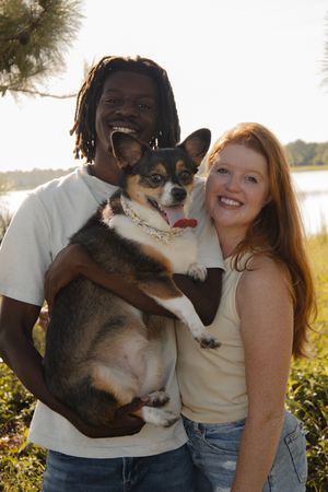 Smiling couple holding a tri-color Corgi outdoors near a lake in golden hour light.