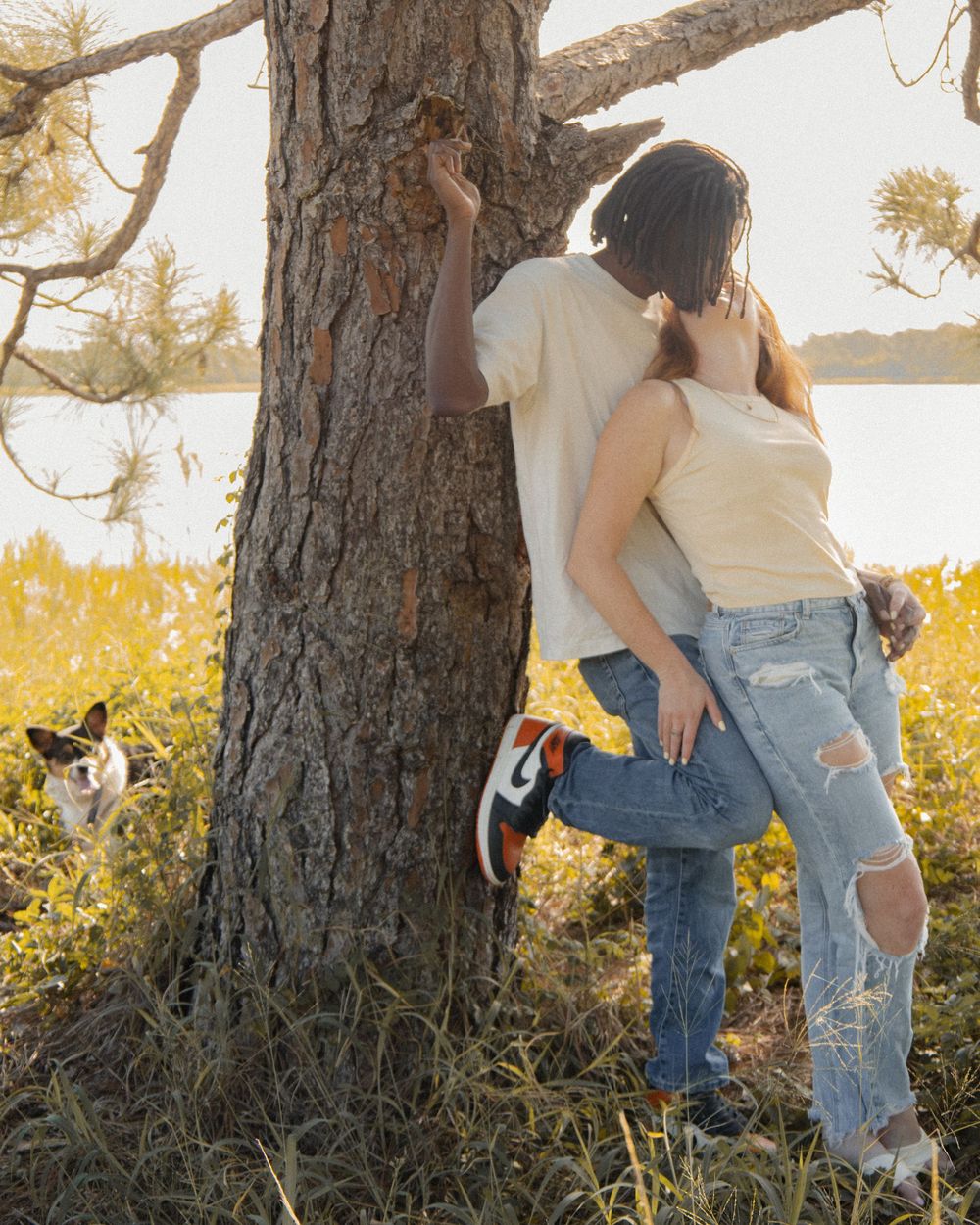 Romantic couple leaning against a pine tree near a lake with a Corgi peeking out beside them.