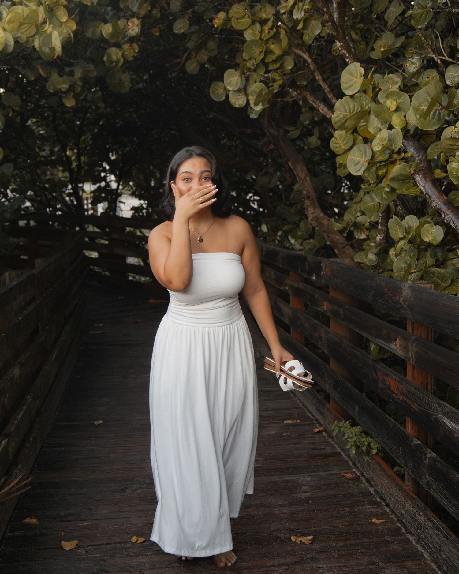 Woman in white strapless maxi dress standing on wooden boardwalk surrounded by lush green trees.
