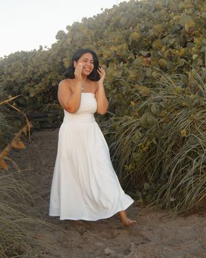 Woman in white strapless maxi dress smiling barefoot on sandy path surrounded by lush green coastal vegetation.