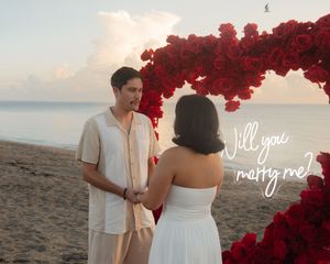 Romantic beach proposal with red rose arch, couple holding hands at sunset with Will you marry me text.