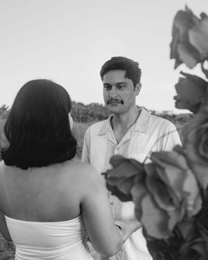 Black and white photo of a groom gazing at his bride during an outdoor wedding ceremony with flowers in the foreground.