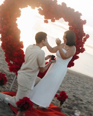 Man proposes to woman on beach under red rose heart arch at sunset, romantic beach proposal scene.