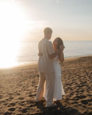 Couple embracing on a sandy beach at golden sunset, wearing white outfits, standing by the ocean shore.