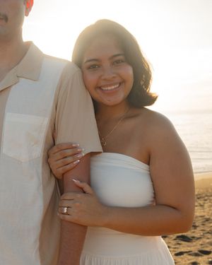 A smiling woman in a white strapless dress embraces a man on a sunny beach at golden hour.