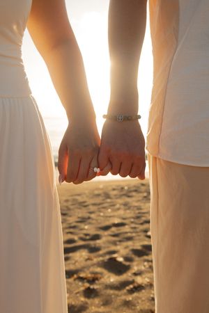 Couple holding hands at sunset on beach, woman wearing engagement ring and bracelet, romantic golden hour.