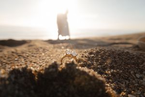 Close-up of a diamond engagement ring resting in beach sand with a blurred figure standing in sunlight behind it.