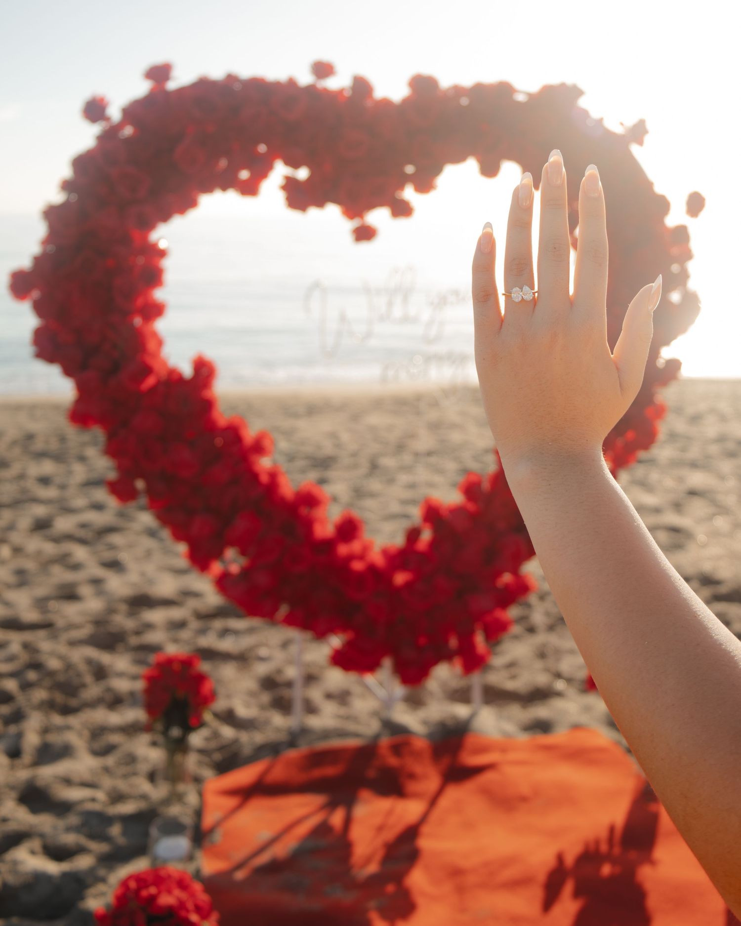 A hand with an engagement ring raised in front of a red rose petal heart on a sandy beach at sunset.