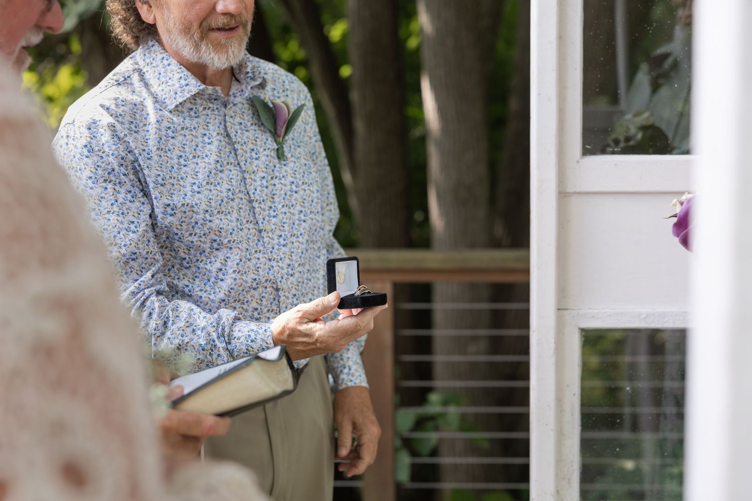 Man in floral shirt holding ring box at outdoor wedding ceremony.