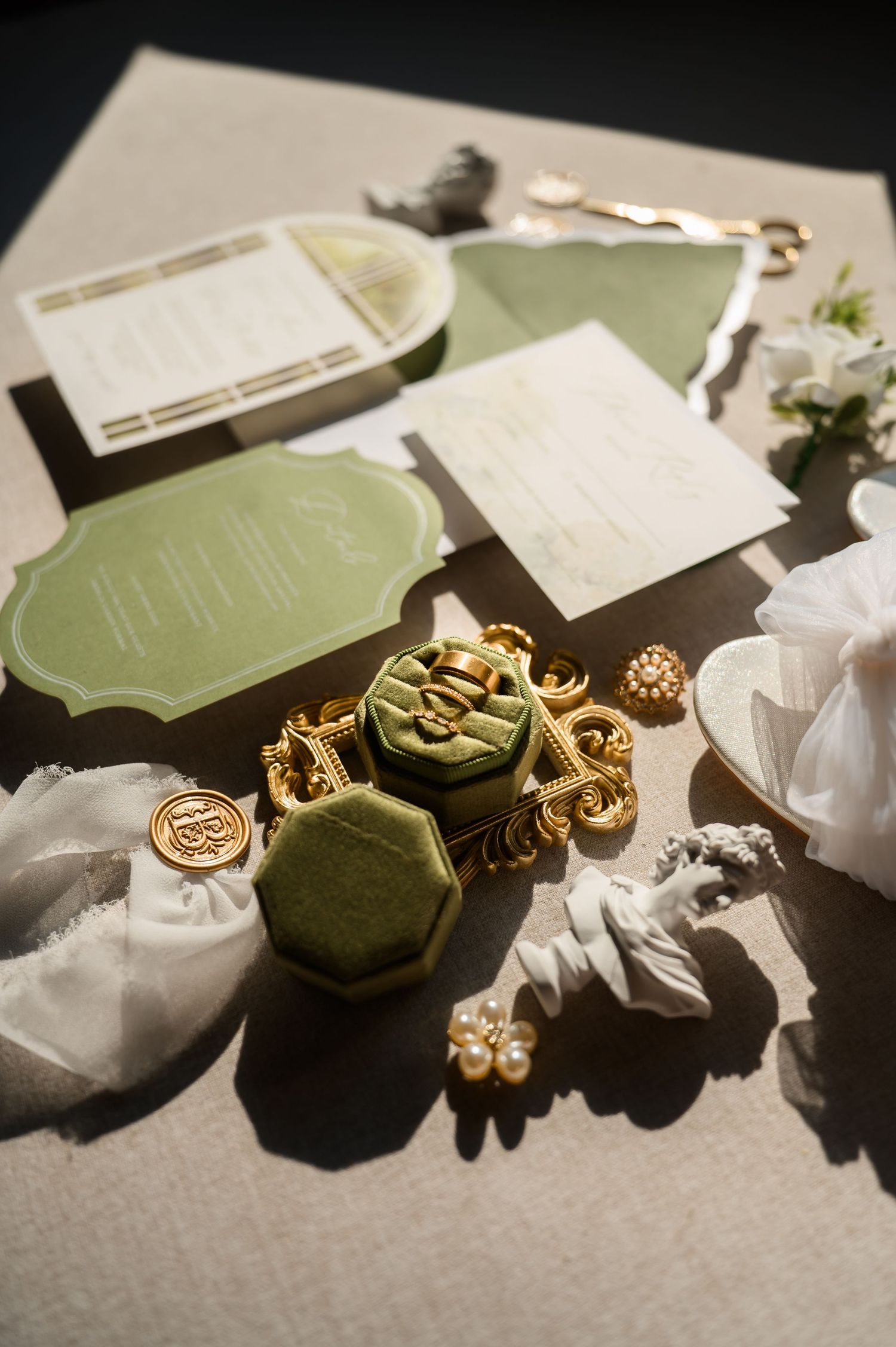 Elegant wedding flat lay with sage green stationery, gold ornate ring box, pearl earrings, and white florals on neutral linen.