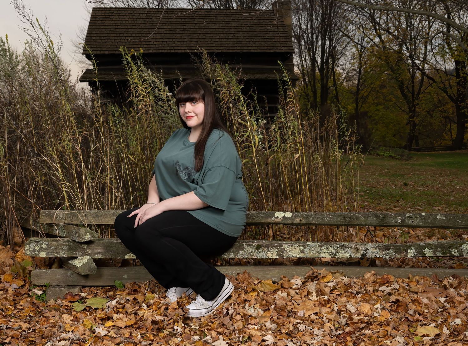 Young woman in green sweatshirt sitting on a bench surrounded by autumn leaves and tall grasses outdoors.