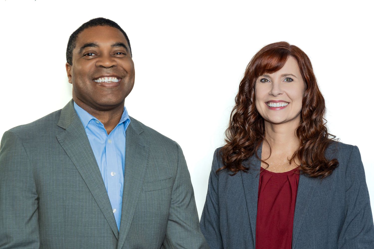 A smiling man in a blue suit and a woman with red hair in a red top pose together against a white background.