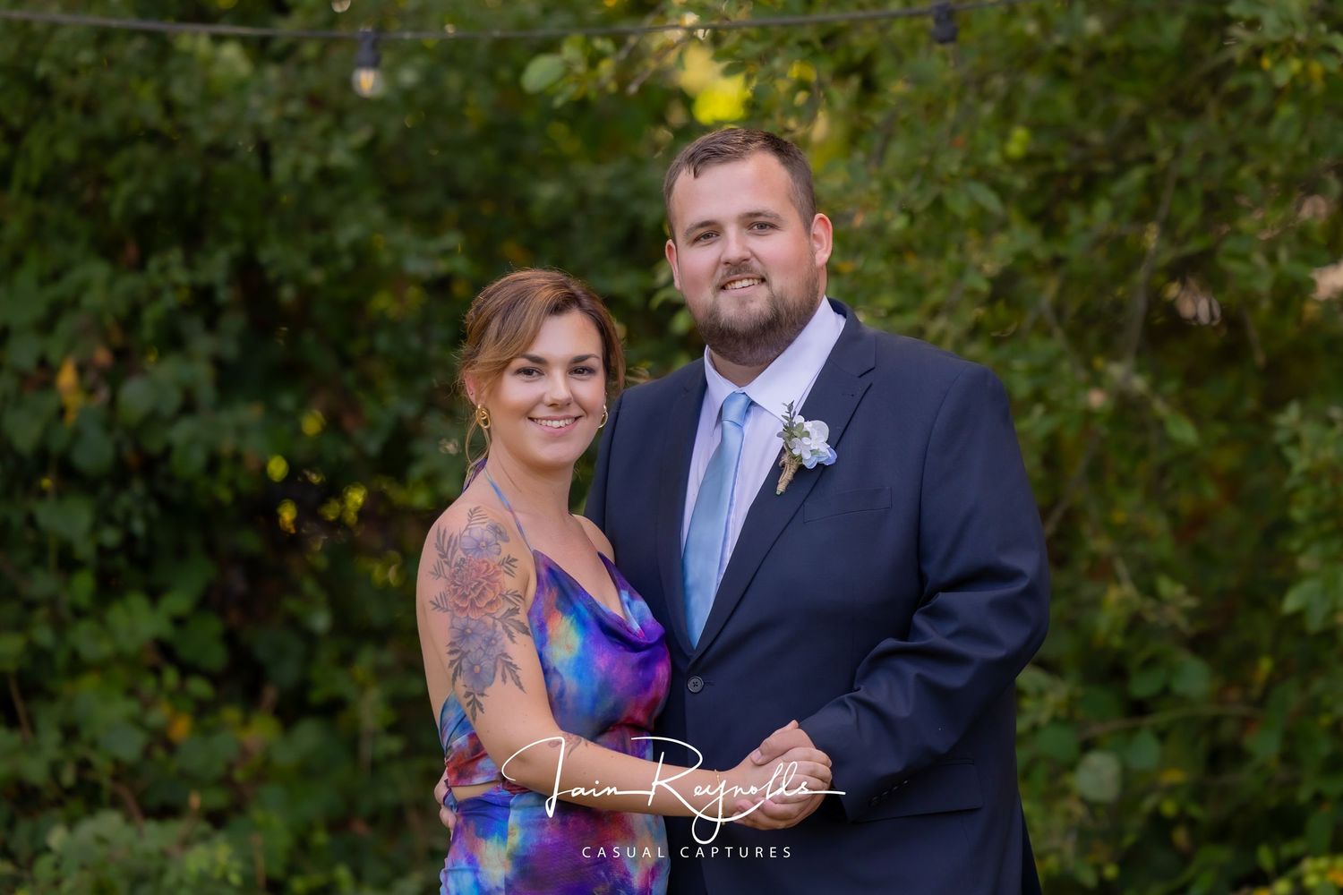 Smiling couple posing outdoors at a wedding, man in navy suit with blue tie, woman in colorful dress, green background. Casual Captures UK