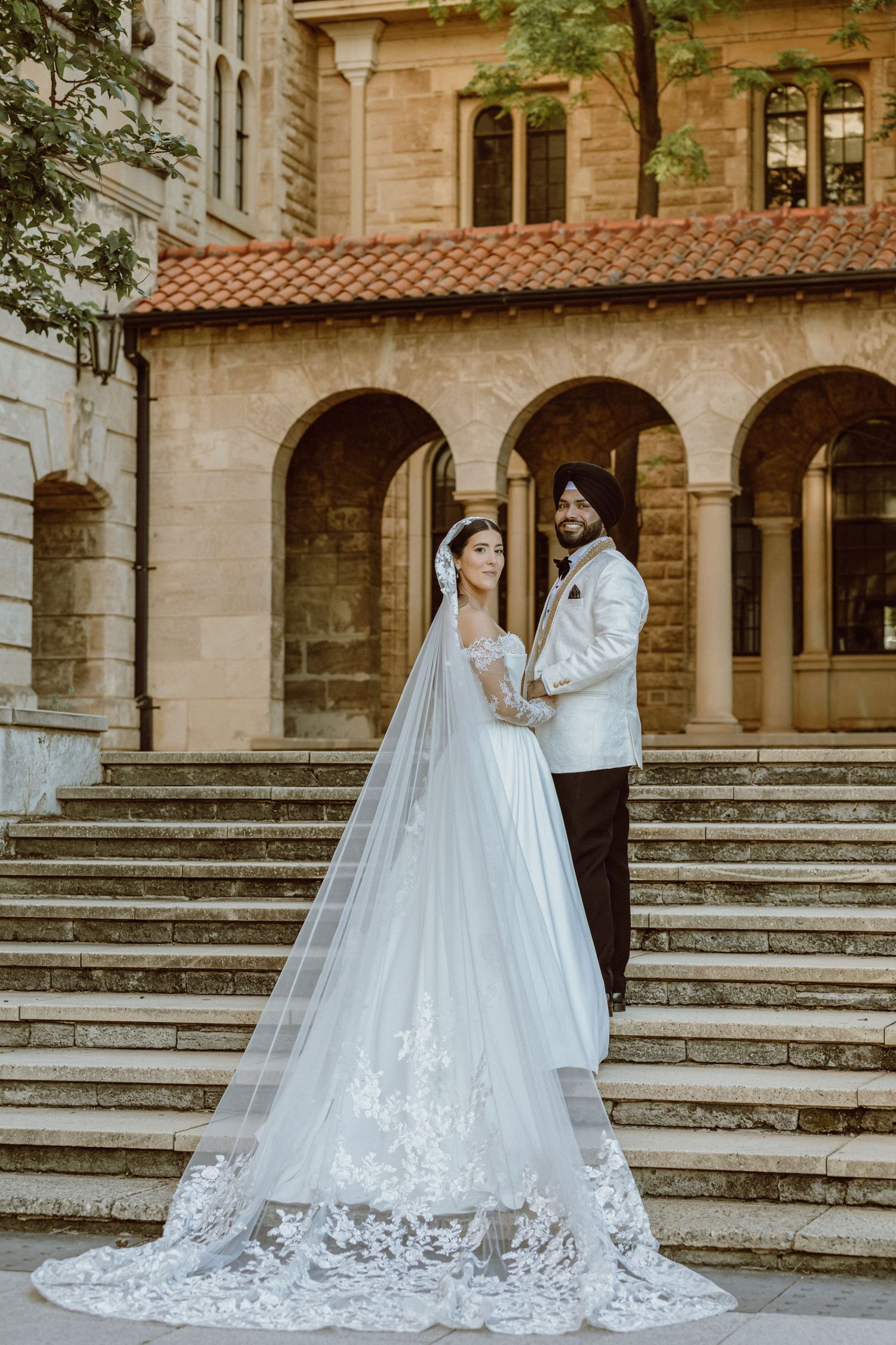 Sikh bride and groom in traditional attire pose beside a vintage white car on their wedding day.