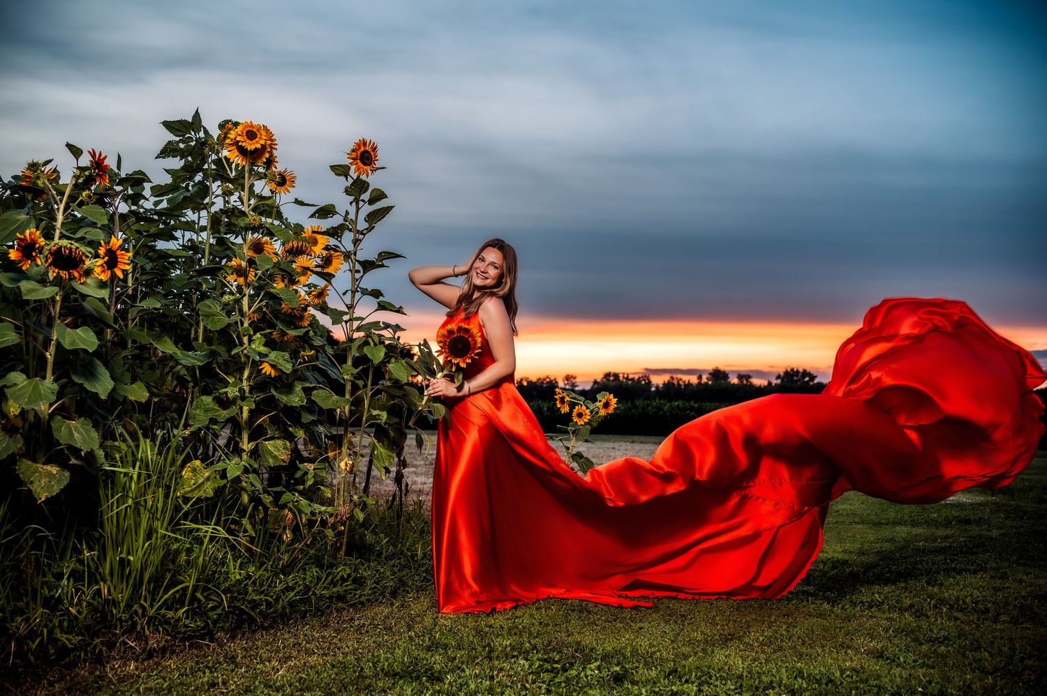 Woman in flowing red gown poses near sunflowers at sunset, fabric billowing dramatically in the wind.