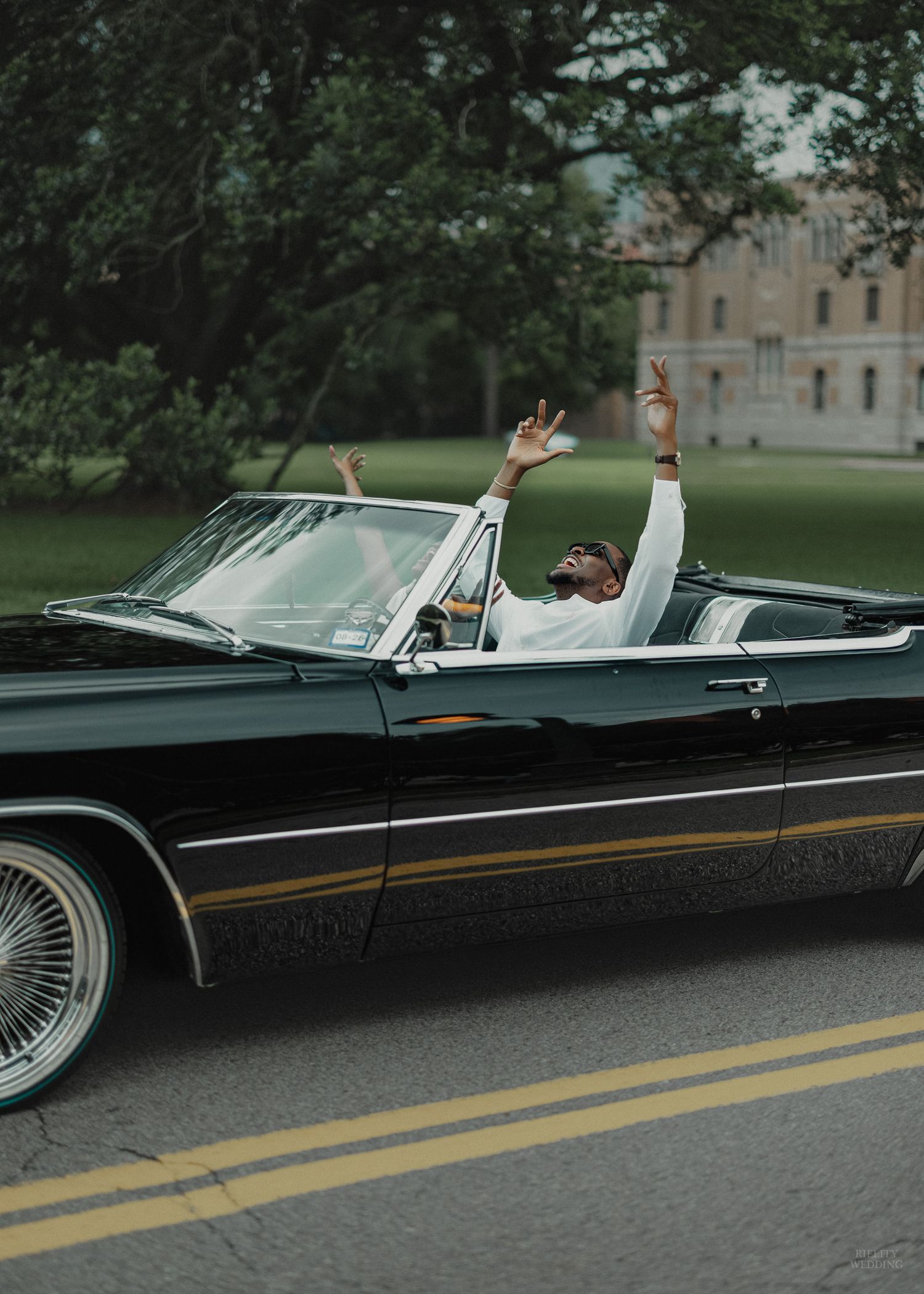 Man in white shirt celebrating with raised hands in a classic black convertible Cadillac driving down a tree-lined road.