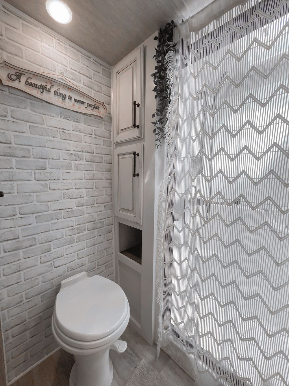 Rustic white brick RV bathroom with white toilet, built-in cabinet storage, and chevron patterned shower curtain.