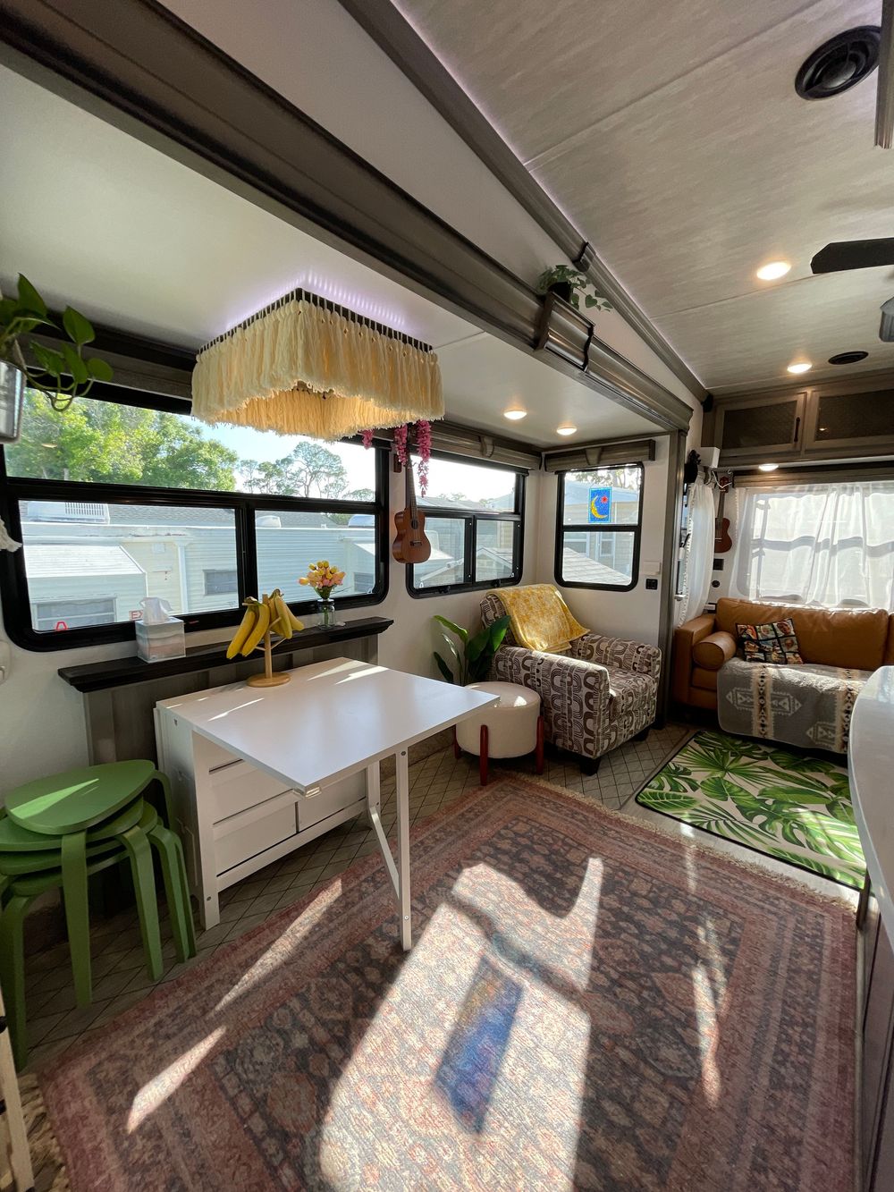 Bright RV dining area with white table, green stools, boho decor, and sunlit living space beyond.