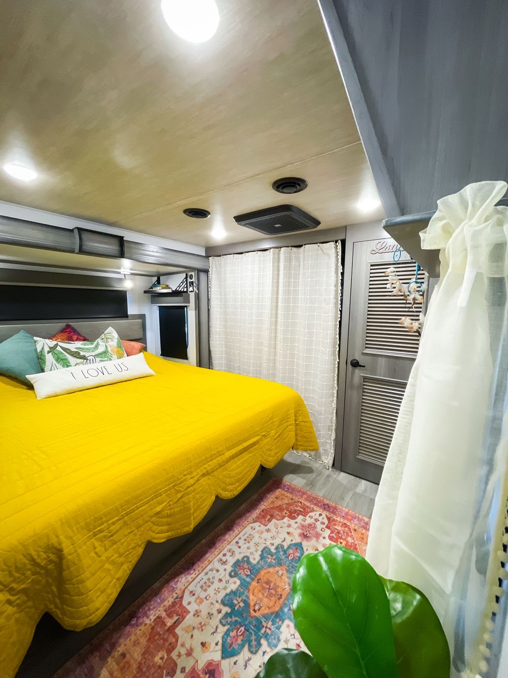 Bright RV master bedroom with bold yellow bedspread, colorful rug, gray ceiling, and sheer white curtains.