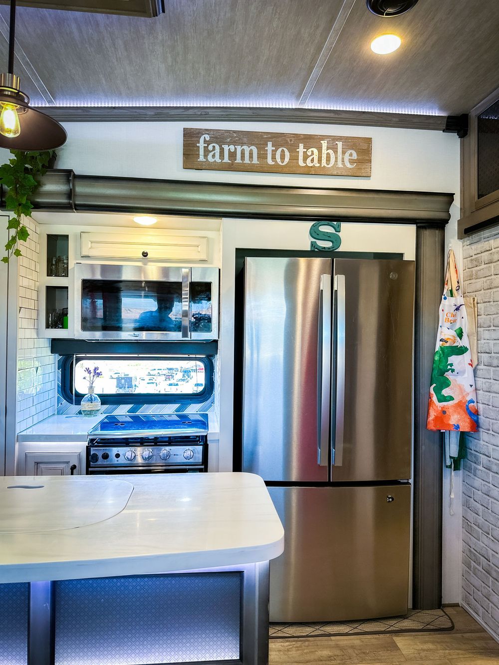Modern RV kitchen featuring stainless steel French door refrigerator, farm to table sign, and brick backsplash.