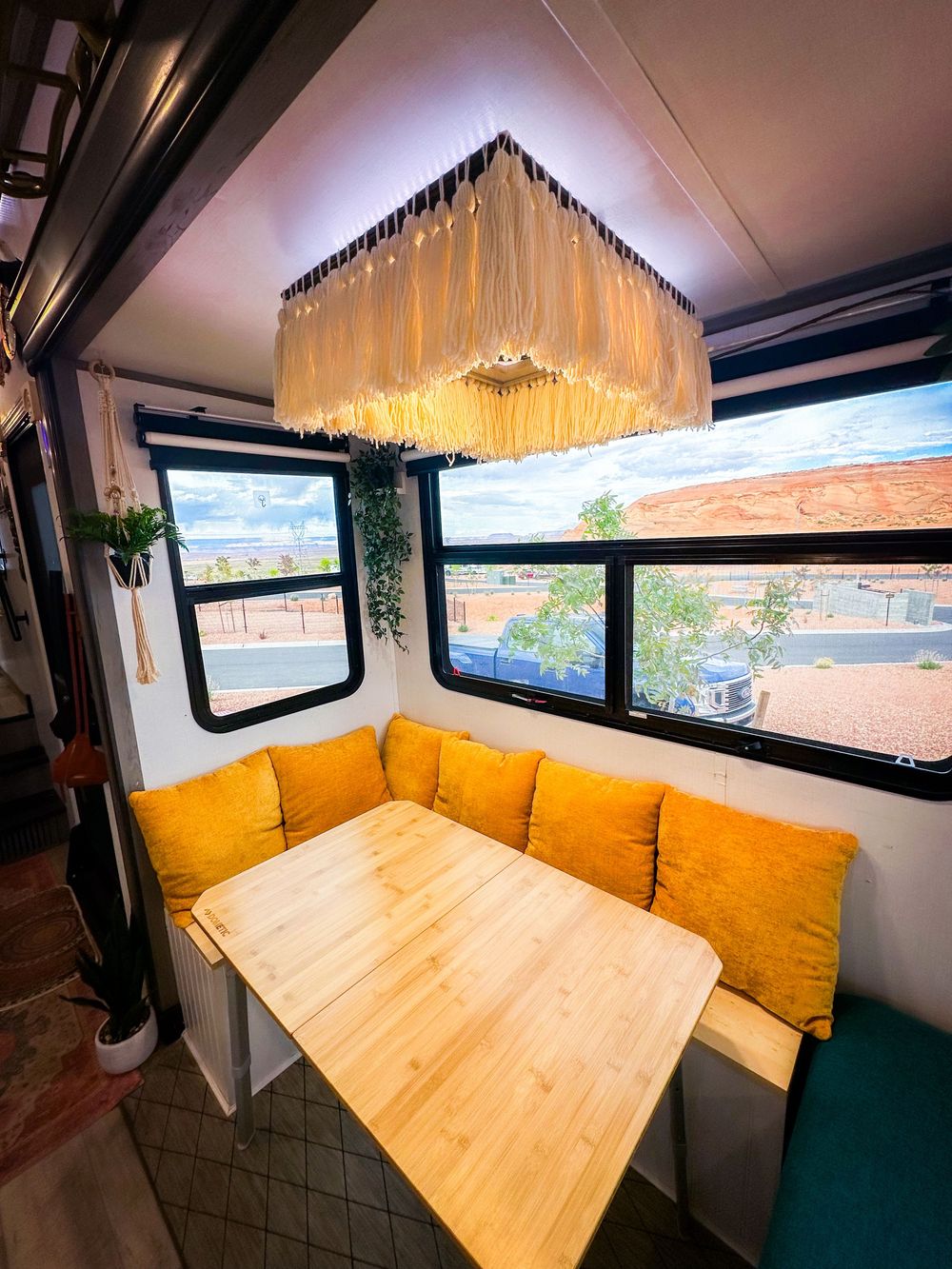 Cozy RV dining nook with golden yellow cushions, wood table, boho fringe chandelier, and scenic window views.