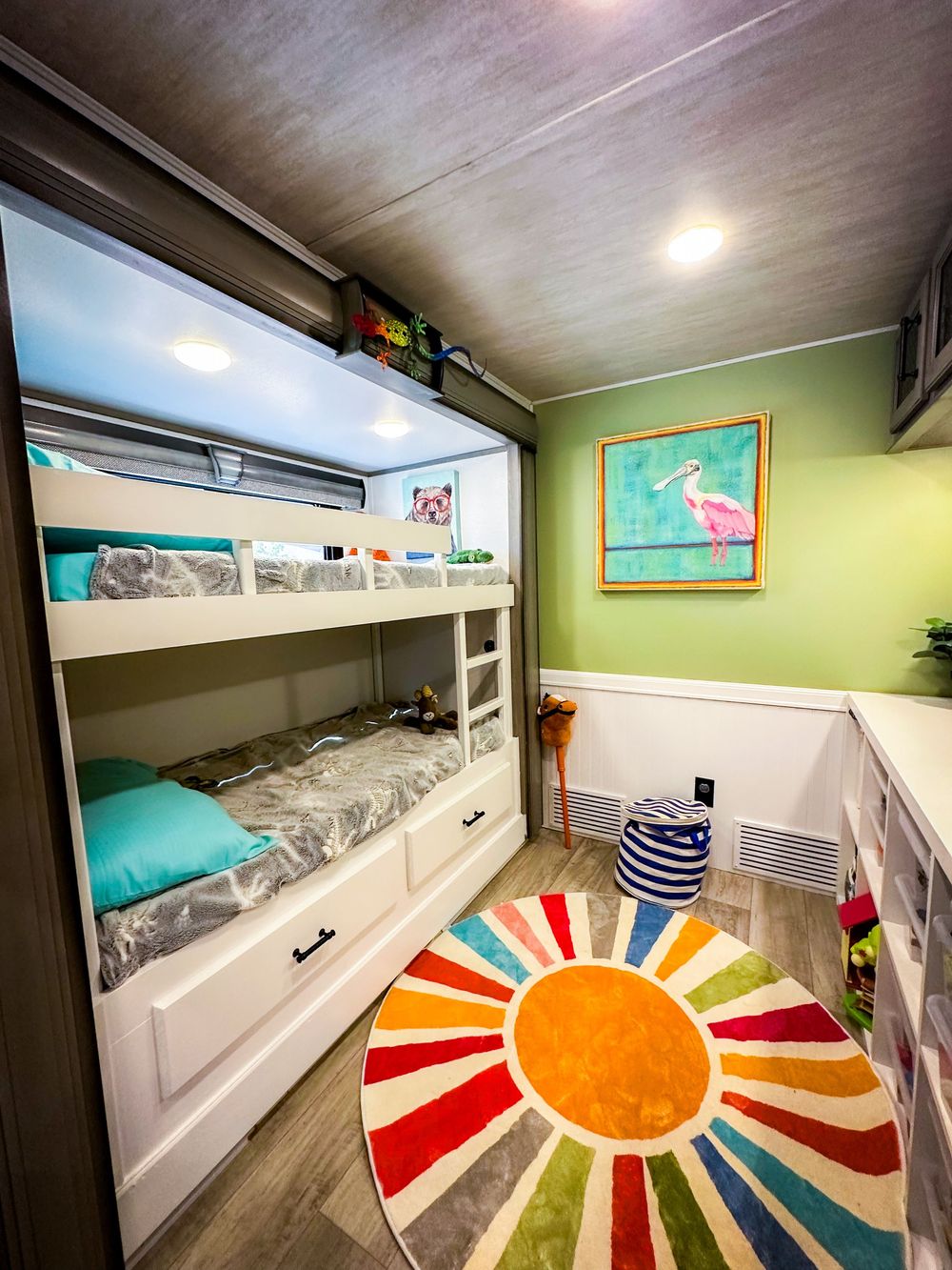 Colorful kids bunk bed room with teal accents, rainbow sun rug, and flamingo wall art in a stylish RV interior.