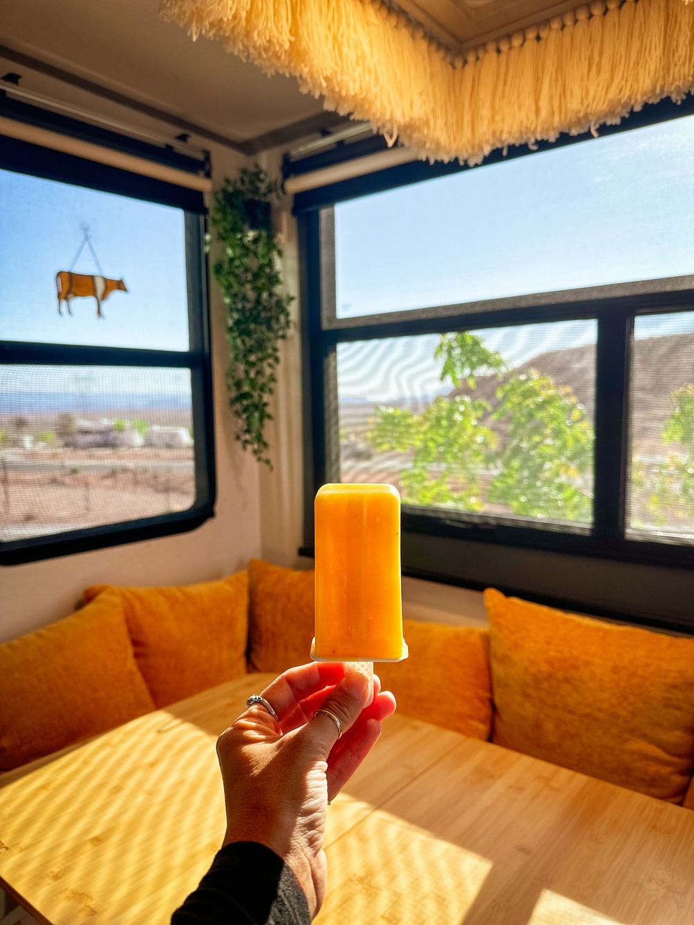 Hand holding orange popsicle in front of sunny RV dinette with mustard cushions.
