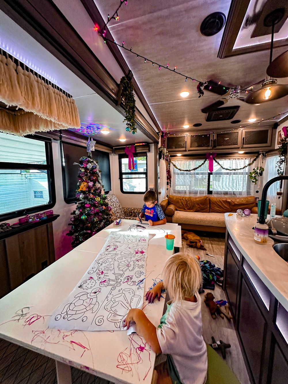 Children coloring a large paper on a table inside a festively decorated RV with Christmas tree and string lights.