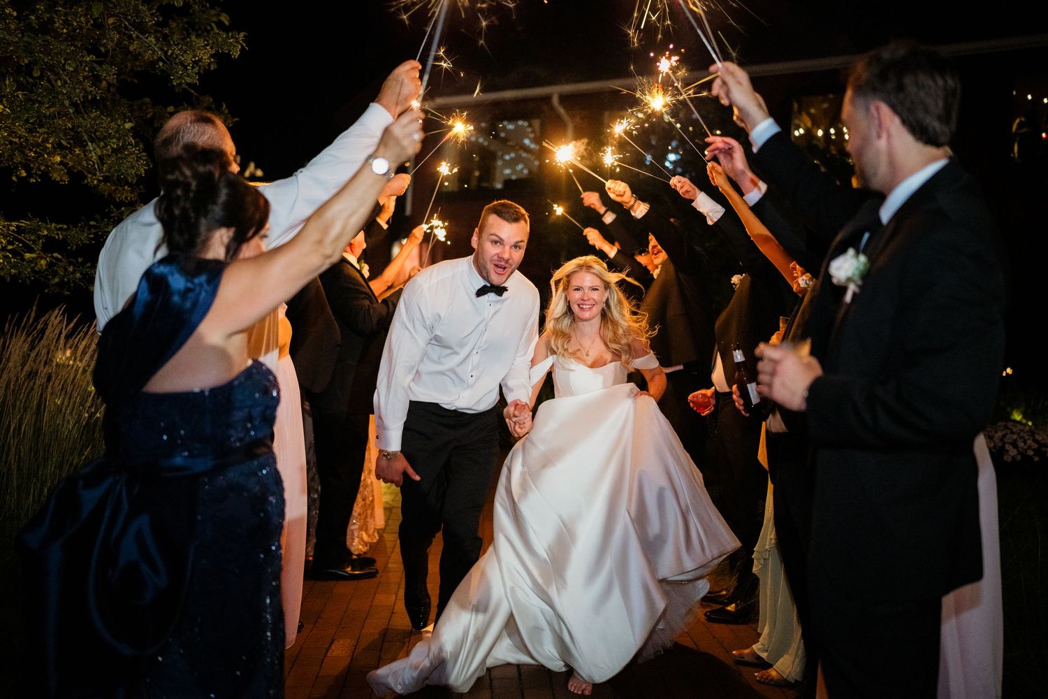 Bride and groom walk through sparkler exit surrounded by cheering wedding guests at night.