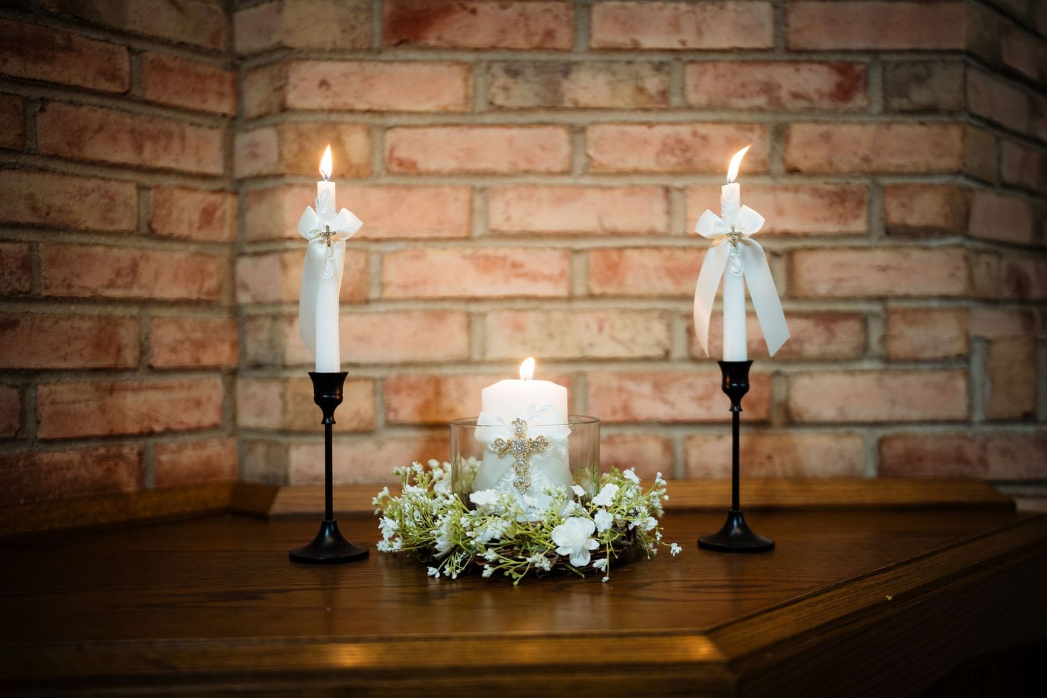 Three white candles with a floral wreath and cross on a wooden surface against a brick wall.