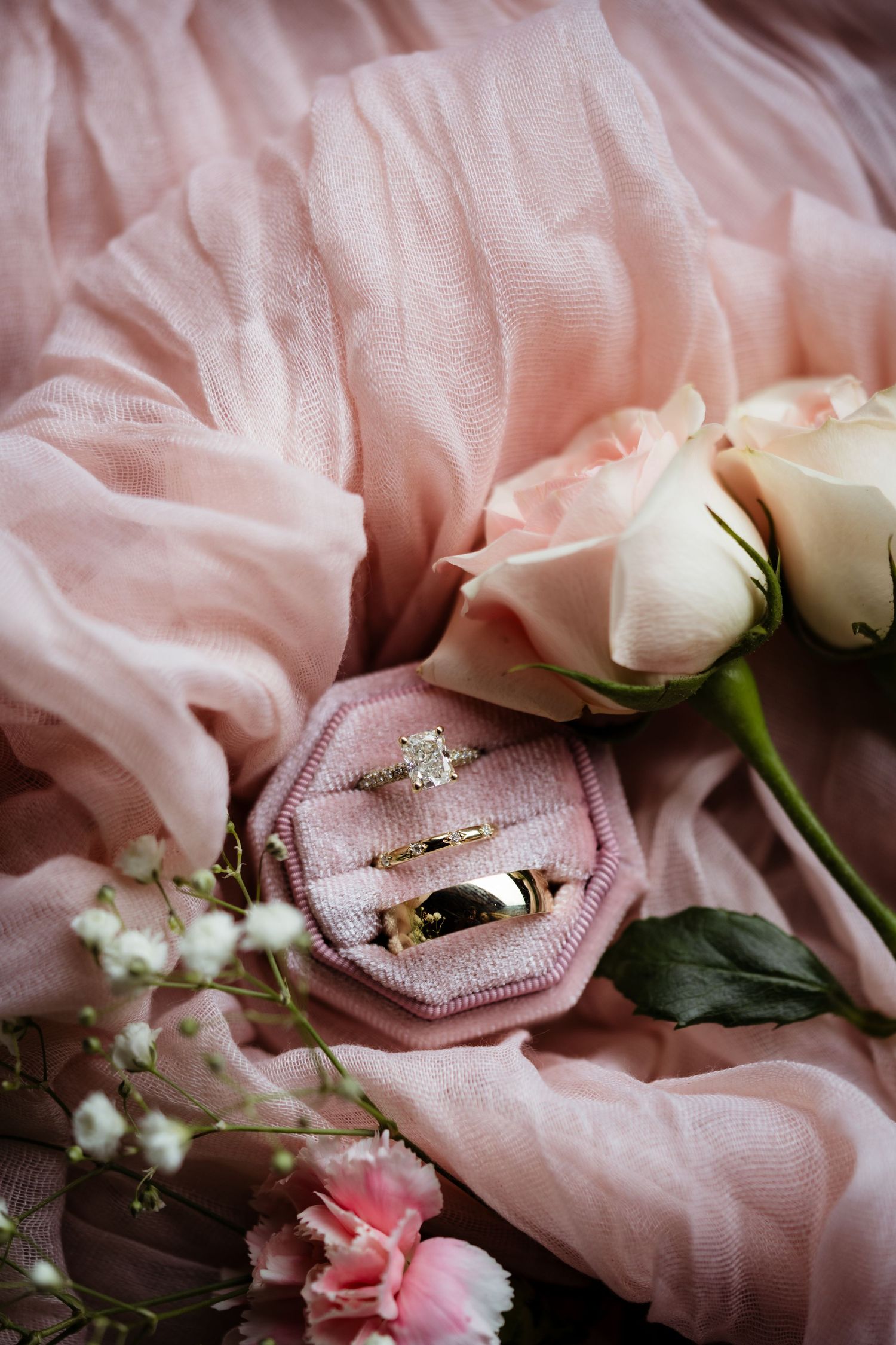Gold wedding rings in a pink velvet box surrounded by roses and baby's breath on soft pink fabric.