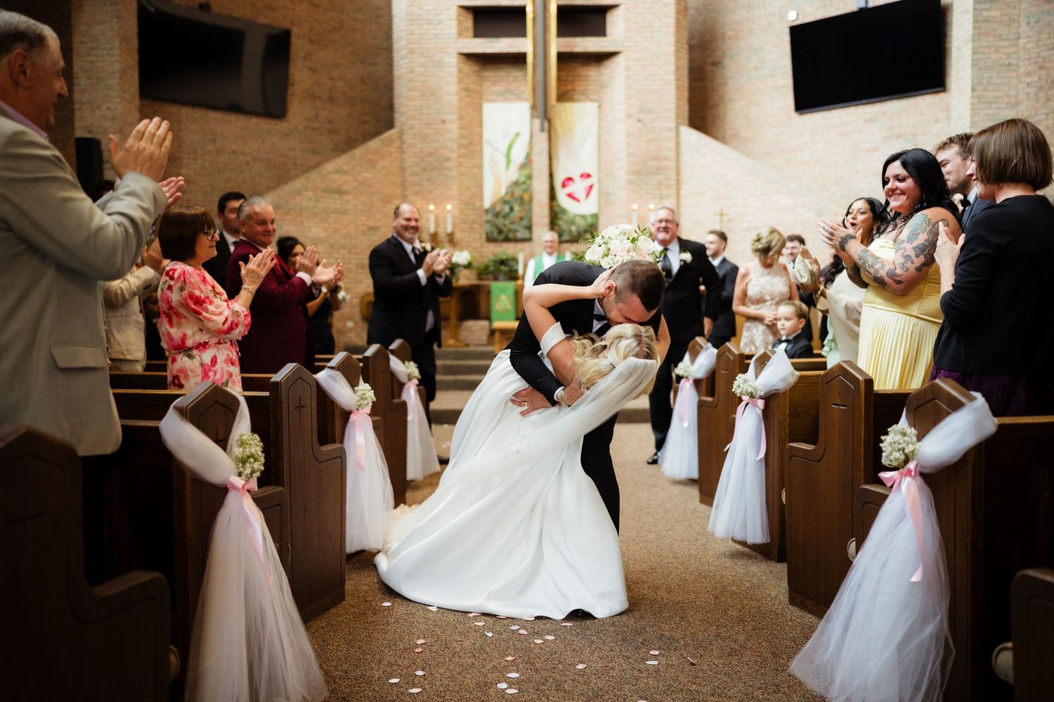 Bride and groom share a romantic dip kiss in church aisle surrounded by applauding wedding guests.