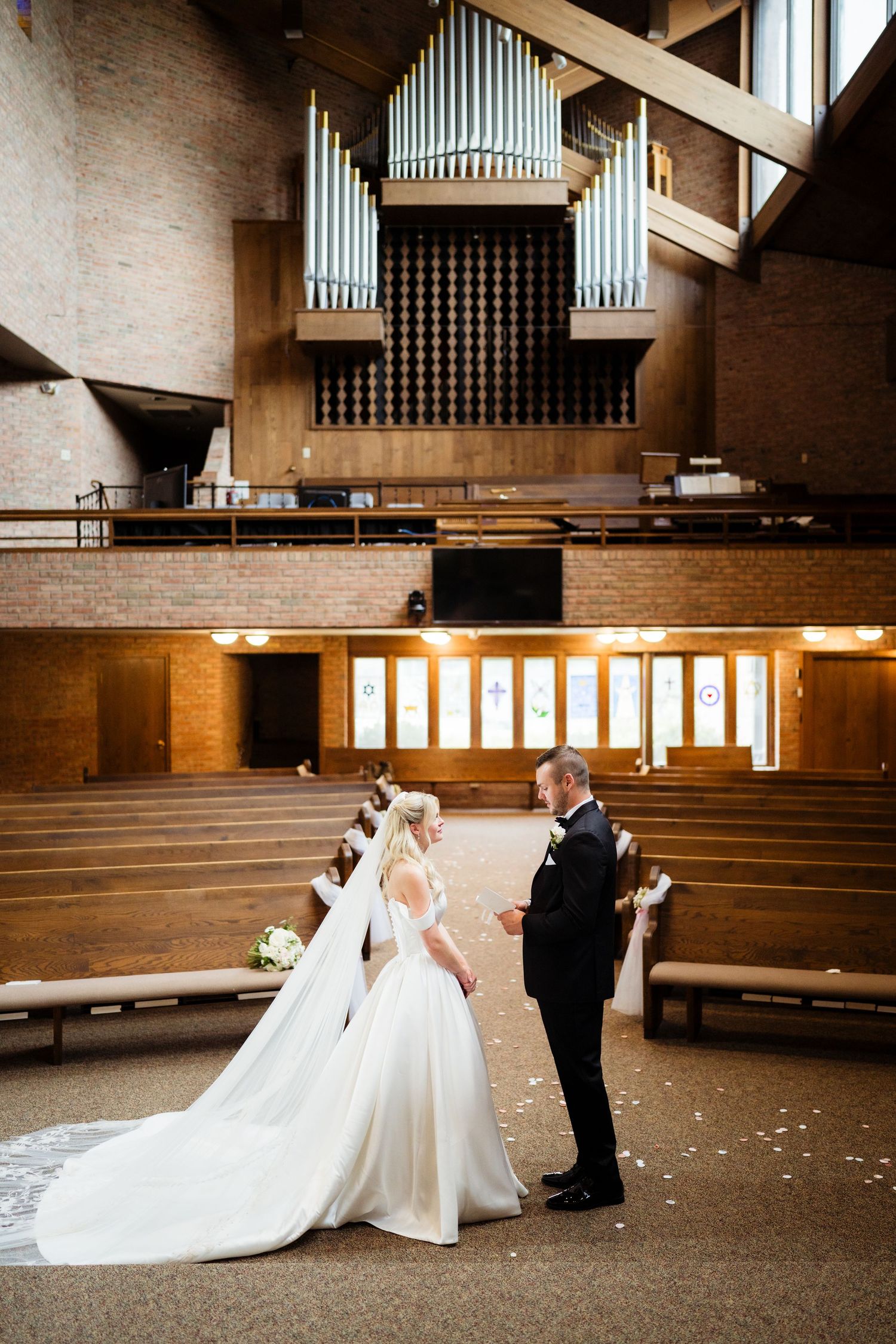 Bride and groom exchange vows at church altar with grand pipe organ backdrop and rose petal aisle.