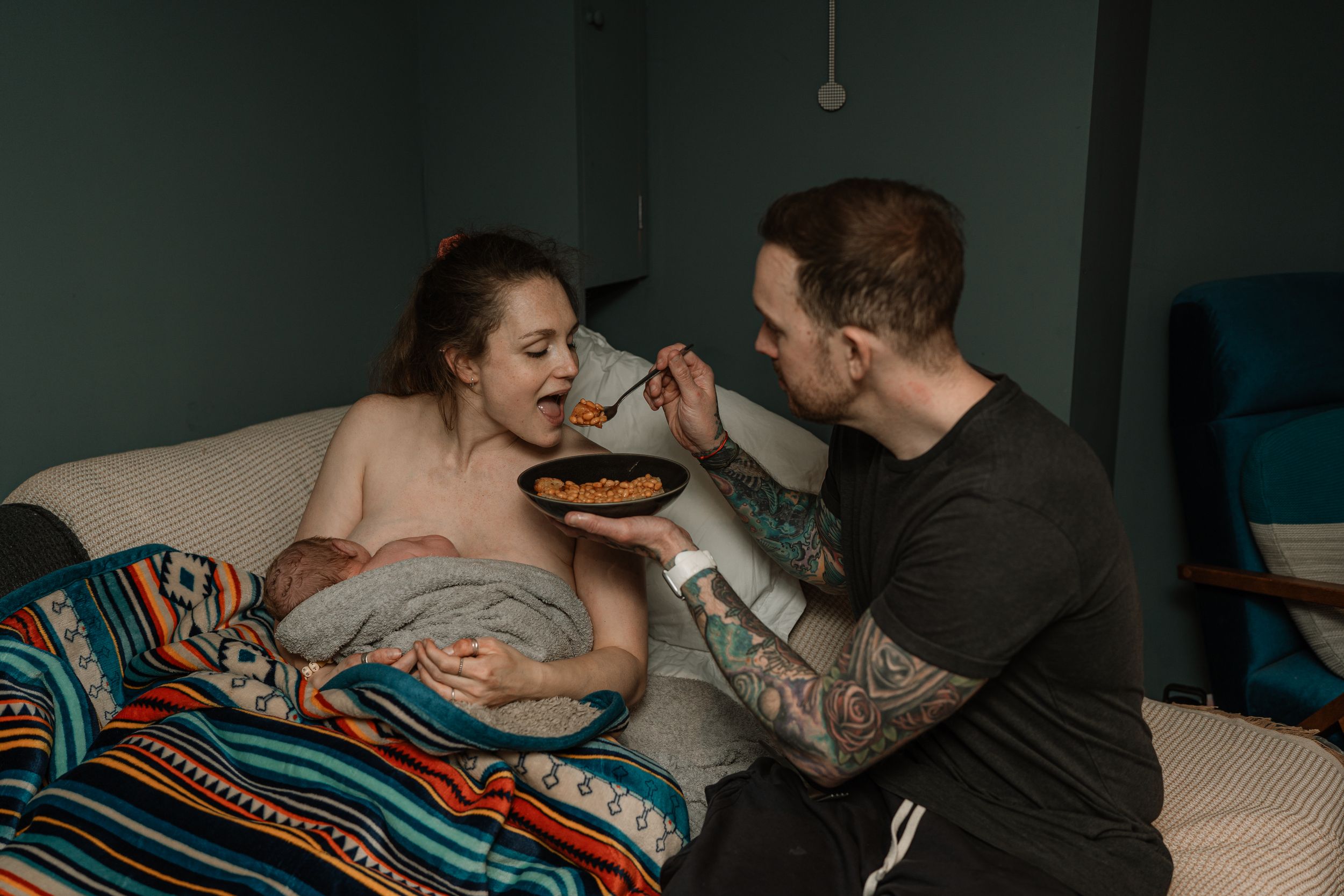 Birth partner feeding a woman resting in bed, sharing an intimate postpartum moment at home.